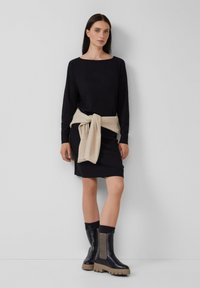 Vestido negro de manga larga con una silueta ajustada, combinado con un suéter de punto crema atado en la cintura y botas negras hasta los tobillos con acentos beige.