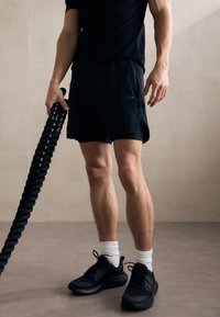 Pantalones cortos deportivos negros con un logo sutil, combinados con una camiseta negra y zapatillas negras. Una cuerda de ejercicio gruesa y retorcida sostenida en una mano.