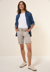 Camicia di jeans sopra una maglietta bianca, abbinata a pantaloni corti beige chiaro. I pantaloni corti hanno orli arrotolati e vita con cordoncino. Sneakers bianche.