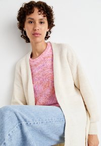 Krämfärgad stickad cardigan med vertikala texturdetailler, lager på en tröja i rosa och orange med prickar. Ljust blå, vida jeans.