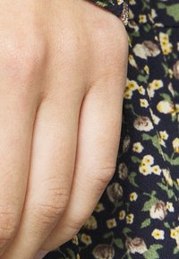 Primo piano di una mano con pelle chiara, caratterizzata da una superficie liscia e vene visibili, poggiata su un tessuto floreale scuro con piccoli fiori gialli e crema.