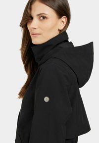 Musta huppari, jossa on mattapintainen viimeistely, vasemmassa hihassa pyöreä logo-patch ja muotoiltu kaulus. Sujuva rakenne.