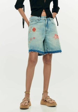 Nő világoskék farmer sortban, színes hímzéssel és rojtos szegéllyel, természetes színű platform szandállal párosítva.