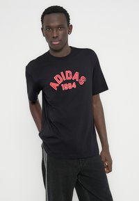 Μαύρο βαμβακερό μπλουζάκι με κόκκινη κεντημένη επιγραφή "ADIDAS 1994" στο στήθος. Το μοντέλο φοράει με σκούρο τζιν.