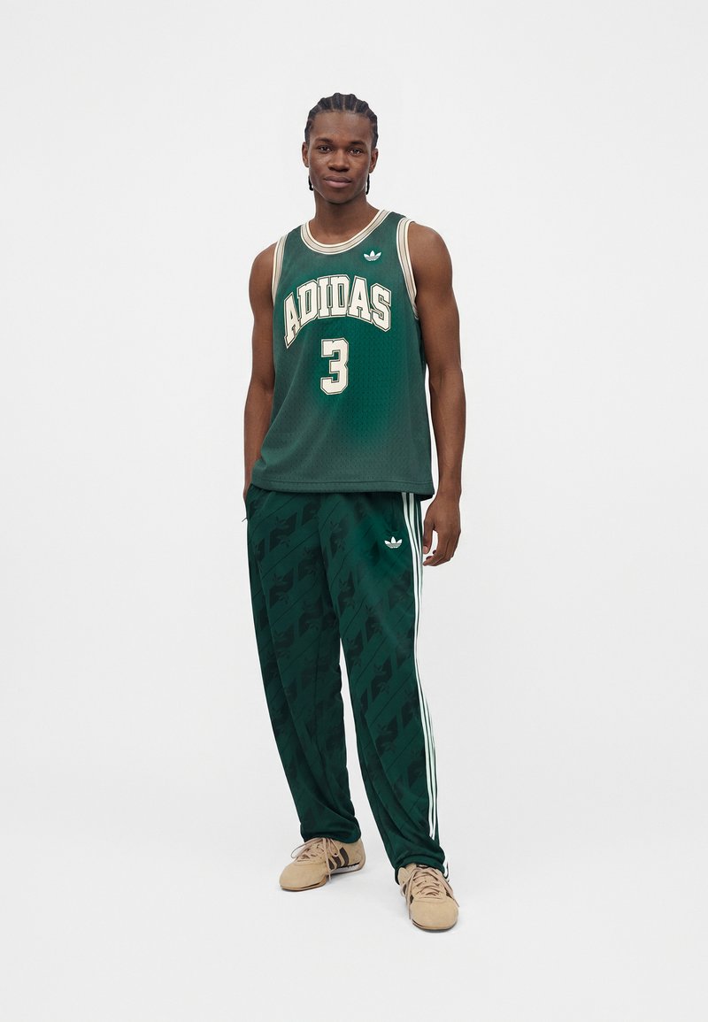 Mann steht und trägt ein dunkelgrünes ärmelloses Adidas-Trikot mit der Nummer 3 und passende grüne Adidas-Trainingshosen mit weißen Streifen.