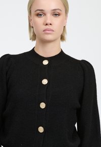 Schwarze Strickjacke mit hohem Kragen, puffigen Ärmeln und fünf goldfarbenen, gerippten Knöpfen auf der Vorderseite. Strukturierter Stoff.