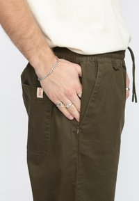 Olijfgroene broek met elastische tailleband, zijzak en een klein label. Het materiaal lijkt soepel met een casual pasvorm.