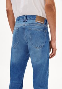 Jean en denim bleu clair avec taille haute, poches arrière et un patch en cuir marron sur la ceinture. Coupe droite classique.
