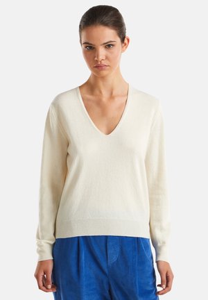 Junge Frau, die einen cremefarbenen V-Ausschnitt-Pullover und leuchtend blaue Cord-Hosen trägt, steht vor einem schlichten weißen Hintergrund.