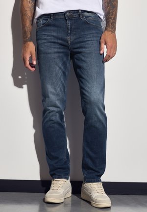 Donkerblauwe denim jeans met een slim fit, voorzien van een klassiek vijf-pocket ontwerp, subtiele vervaging en gestikte details. G gedragen met lichtgekleurde sneakers.