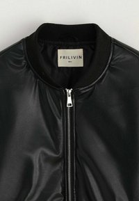 Veste en similicuir noir avec une fermeture éclair frontale argentée et un col en maille côtelée, avec une étiquette de marque blanche cousue à l'intérieur du col portant l'inscription "FRILIVIN."