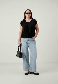 Sieviete, tērpusies melnā kreklā ar savērptu priekšpusi, gaiši zilās džinsās, baltās platformas kurpēs, saulesbrillēs, tur melnu rokassomu, stāv uz vienkāršas fona.