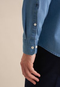 Lichtblauwe denim shirt met een knoopmanchet, voorzien van witte knopen en een zwart geborduurd bloemenmotief nabij de rand van de mouw.