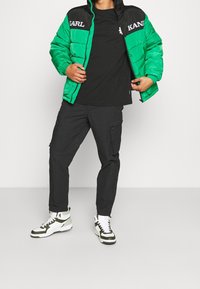 Giubbotto imbottito verde e nero con logo, t-shirt nera, pantaloni cargo scuri e sneakers alte bianche/verde. In piedi su uno sfondo chiaro.
