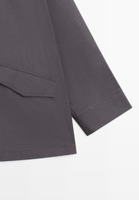 Rękaw ciemnoszarej kurtki z przeszywanym mankietem i detalem klapki na gładkiej powierzchni materiału.