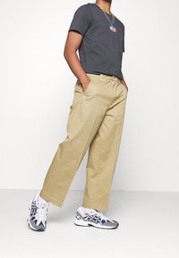 Une personne portant un t-shirt gris avec un logo, vêtue de chinos larges beiges et de baskets grises avec des accents noirs, se tient devant un fond blanc.