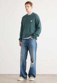 Teal waffle-gebreide trui met ronde hals, gecombineerd met verlichte blauwe jeans met grote gescheurde knieën. Casual outfit met beige schoenen.