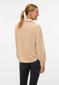 Camicia beige con bottoni, maniche lunghe e colletto. Presenta un tessuto texture e bottoni sui polsini. Indossata con pantaloni neri, vista dal retro.