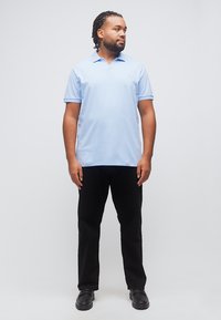 Άνδρας με πλεξούδες, φορούσε γαλάζιο πόλο πουκάμισο, μαύρο παντελόνι και μαύρα παπούτσια, στέκεται και κοιτάζει προς το πλάι με φόντο μια απλή επιφάνεια.