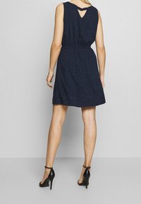 Marinblå ärmlös klänning med insydd midja, har en skärning i ryggen och ett litet prickmönster. Bärs med svarta klacksandaler.