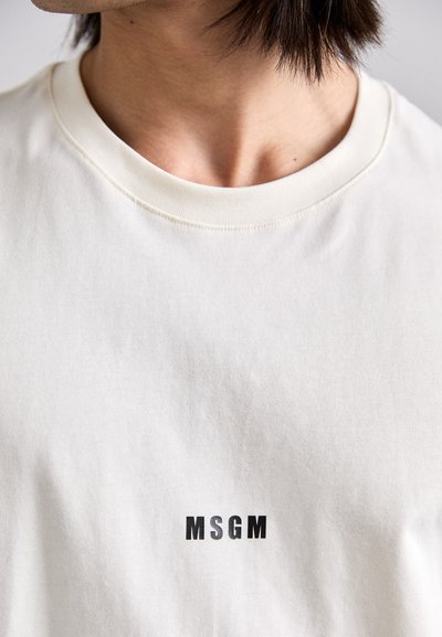Lys beige bomulls t-skjorte med rund hals og en svart "MSGM" logo trykket sentralt. Tøyet har en glatt overflate med en subtil tekstur.