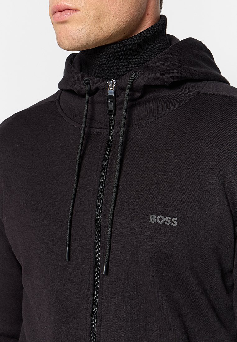 Zwarte zip-up hoodie gemaakt van zachte stof, met een hoge kraag, koordjes en een klein logo op de borst. Gelaagde oppervlakte.