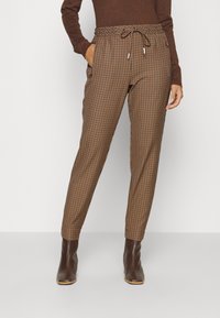 Pantalon marron à carreaux avec une ceinture élastique et un cordon de serrage, doté de poches latérales et de jambes fuselées. Porté avec des bottines marron.
