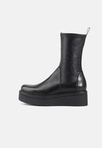 Botas de tobillo de cuero negro con punta redonda, caña elástica y suela de plataforma negra gruesa y texturizada. Cuenta con una lengüeta trasera.