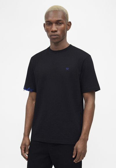 Άνδρας με κοντά λευκασμένα μαλλιά που φοράει μαύρο κοντομάνικο μπλουζάκι με μικρό μπλε λογότυπο με σταυρωμένους σφυριά στο στήθος και μπλε γράμματα στο μανίκι.