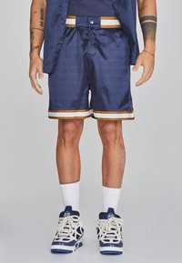 Marineblaues Shorts aus strukturiertem Stoff, mit einem gold-weißen gestreiften Saum, kombiniert mit weißen Knöchelsocken und marineblauen Sportschuhen.