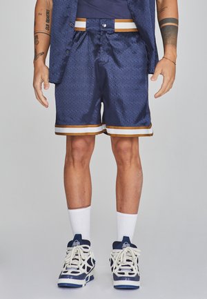 Marineblaues Shorts aus strukturiertem Stoff, mit einem gold-weißen gestreiften Saum, kombiniert mit weißen Knöchelsocken und marineblauen Sportschuhen.