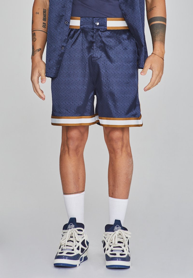 Marineblaues Shorts aus strukturiertem Stoff, mit einem gold-weißen gestreiften Saum, kombiniert mit weißen Knöchelsocken und marineblauen Sportschuhen.
