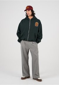 Grønn zip-up hettejakke med frontgrafikk og to lommer, kombinert med lysegrå plisserte bukser og brune sko med hvite detaljer. Brun caps.