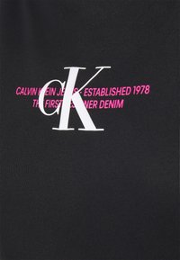 Czarna tkanina z dużymi białymi literami "cK" oraz różowym napisem "Calvin Klein Jeans - Założono w 1978, pierwsze denimowe ubrania projektanta."