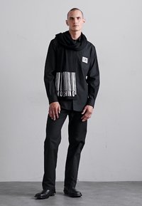 Camisa preta com um gráfico de código de barras na parte inferior, colarinho dobrado e bolso frontal. Usada com calças pretas e sapatos de pele.