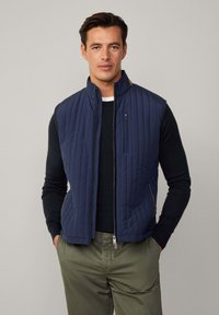 Gilet matelassé marine avec col haut, fermeture éclair sur le devant et poches latérales, porté sur une chemise noire à manches longues, associé à un pantalon vert olive.