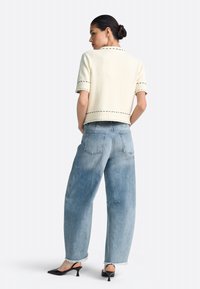 Crèmekleurige trui met korte mouwen en een gestructureerde stof met contrasterende stiksels, gecombineerd met lichtblauwe wijde denim jeans en zwarte mules met hak.