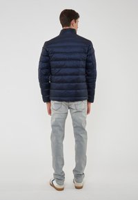 Uomo che indossa una giacca trapuntata blu navy, jeans grigi chiari e scarpe da ginnastica bianche, in piedi con le spalle rivolte verso un fondo bianco semplice.