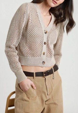 Donna che indossa un cardigan corto beige a maglia aperta con bottoni, infilato in pantaloni color camoscio a vita alta con cintura nera e portachiavi a forma di stella.