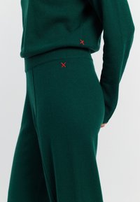 Groene gebreide lounge-set met een losse top en toelopende broeken. Opvallende rode "X"-stiksels op beide stukken. Zachte textuur met ribbelmanchetten.