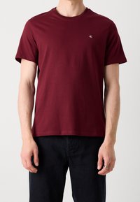 Burgundské bavlněné tričko s kulatým výstřihem a krátkými rukávy; má malý detail loga na levém prsu; hladká textura.