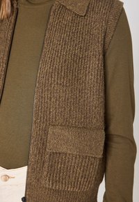 Gilet di maglia marrone con texture a coste, dotato di colletto e tasca frontale. Indossato sopra una maglietta verde a maniche lunghe. Chiusura con cerniera sul lato.