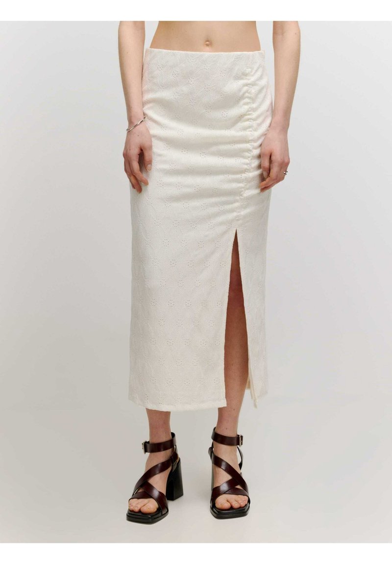 Femme portant une jupe midi blanche avec une fente à l’avant et des sandales à talons hauts marron foncé avec des brides à la cheville sur un fond uni.