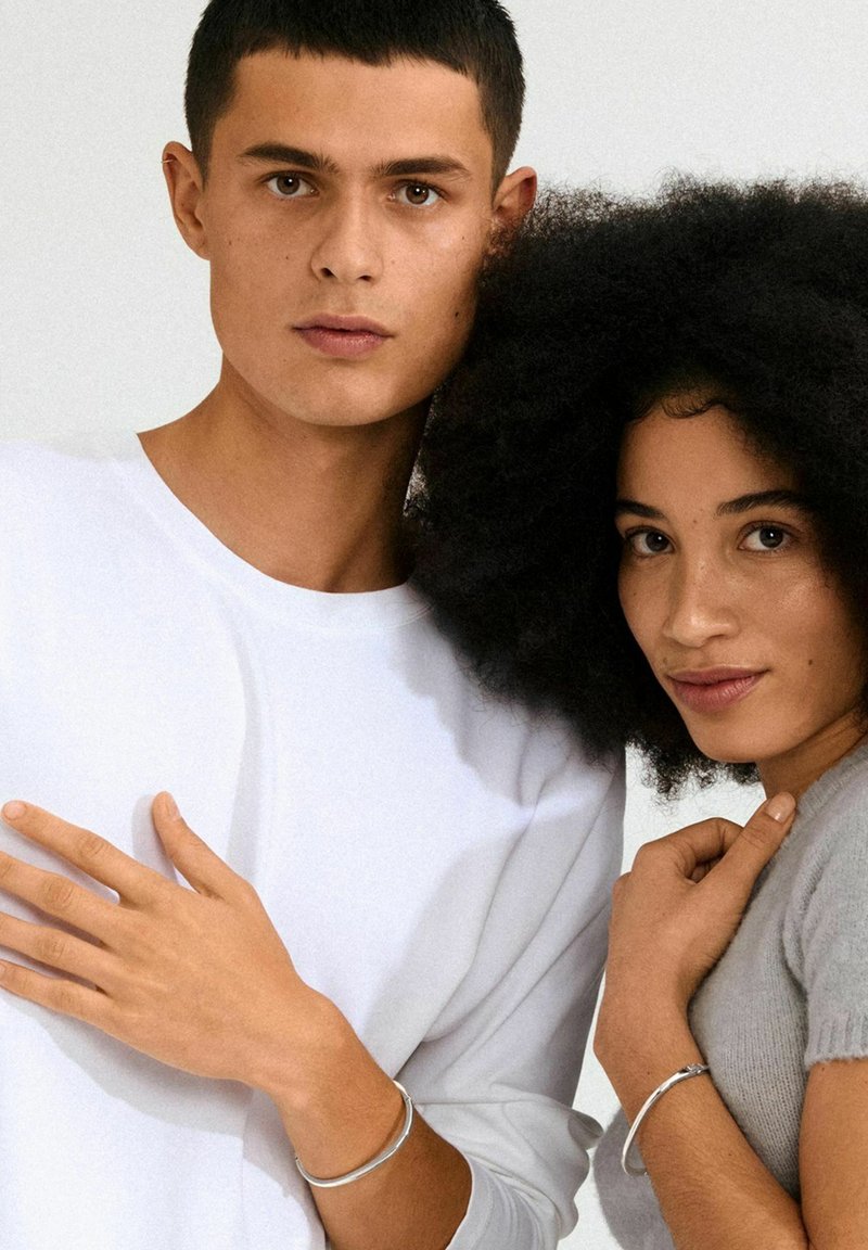 Pair of models wearing simple white and gray clothing. Both have delicate silver bracelets. Skin tones vary; hair textures are smooth and curly.