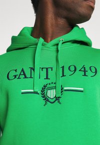 Felpa verde in cotone con cappuccio e cordino, caratterizzata da testo ricamato in blu navy "GANT 1949" e un design con uno stemma decorato con stelle e allori.