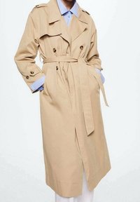 Trench-coat beige avec une taille ceinturée, un devant croisé, des épaulettes et un tissu lisse et léger. Manches longues avec poignets à boutons.