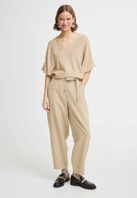 Pull beige à manches courtes et décolleté en V associé à un pantalon beige taille haute, avec un lien à la taille, et des mocassins noirs avec des accents dorés.