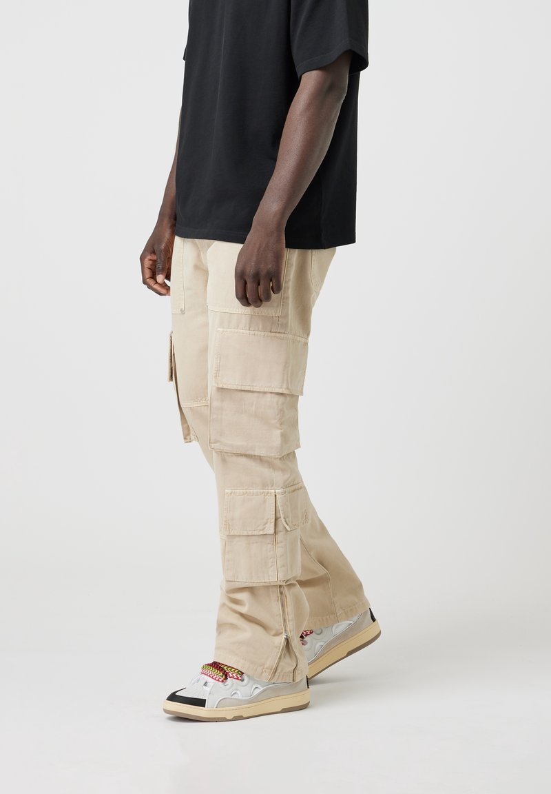 Personne portant un pantalon cargo beige avec plusieurs poches, un t-shirt noir et des baskets blanches avec des lacets rouges et jaunes, debout de profil.