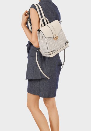 Donna che indossa un abbigliamento senza maniche blu, porta uno zaino bianco con motivo, con cinghie di cuoio e fibbia color oro, appoggiato su una spalla.