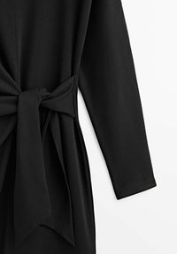Robe portefeuille noire à manches longues, dotée d'une ceinture nouée à la taille, d'une texture lisse et d'un design épuré.